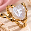 A gold ring featuring a large, lustrous white baroque pearl shaped like a heart, set in a wavy, organic band, displayed on a textured cream surface with soft pink fabric in the background.