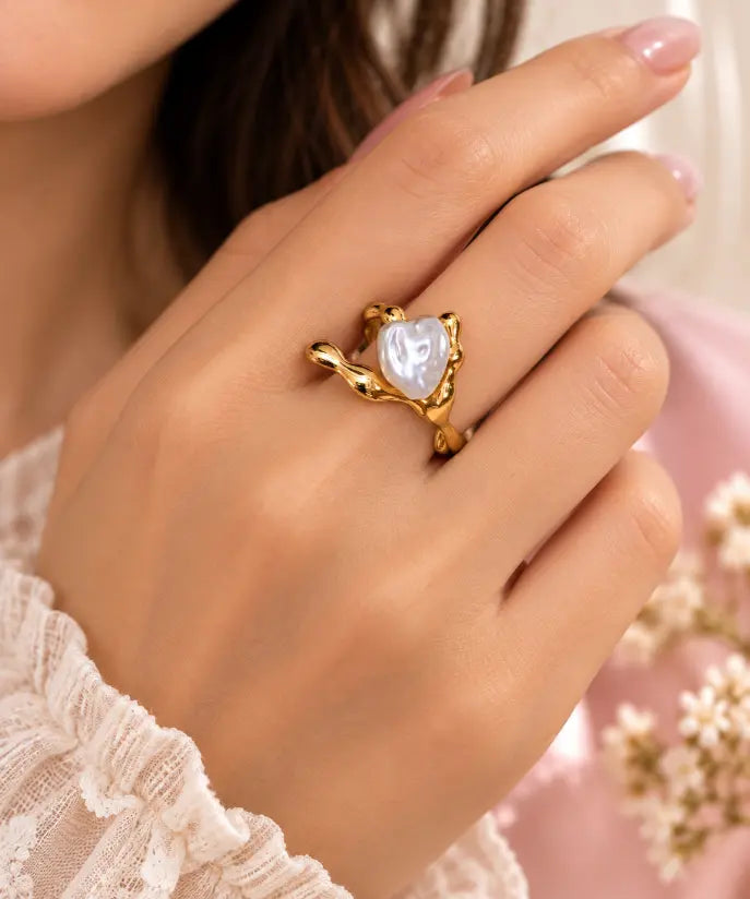 A close-up lifestyle shot of a person's hand wearing a gold ring with a heart-shaped white pearl in a branch setting, against a soft pink background with blurred flowers.