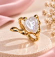 A gold ring with a wavy, organic-shaped setting holding a lustrous white heart-shaped pearl, displayed on a round beige pedestal with soft pink fabric and dried flowers in the background.
