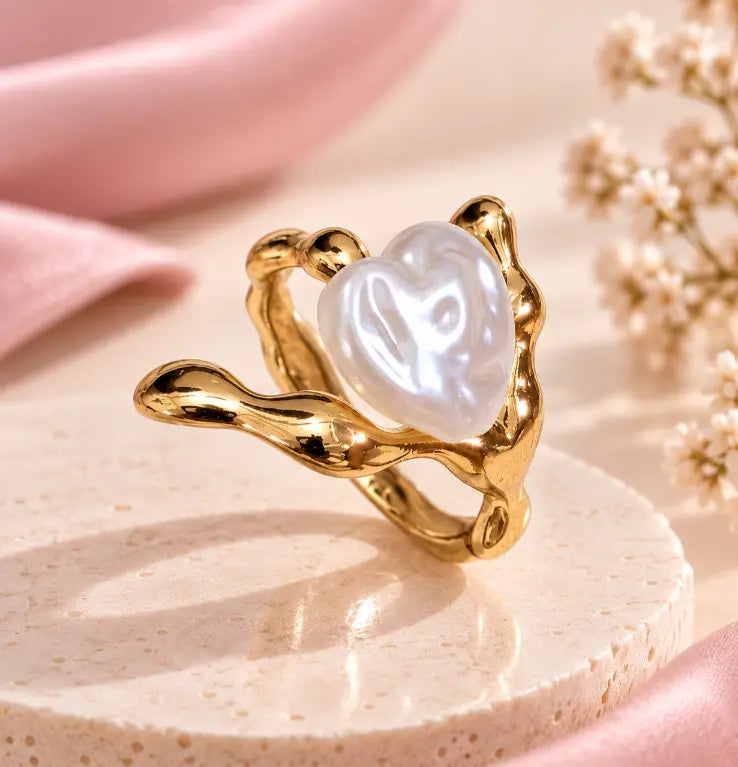 A gold ring with a wavy, organic-shaped setting holding a lustrous white heart-shaped pearl, displayed on a round beige pedestal with soft pink fabric and dried flowers in the background.