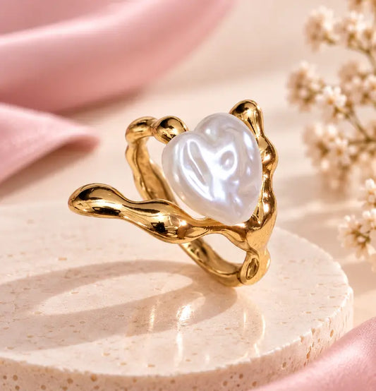 A gold ring with a wavy, organic-shaped setting holding a lustrous white heart-shaped pearl, displayed on a round beige pedestal with soft pink fabric and dried flowers in the background.