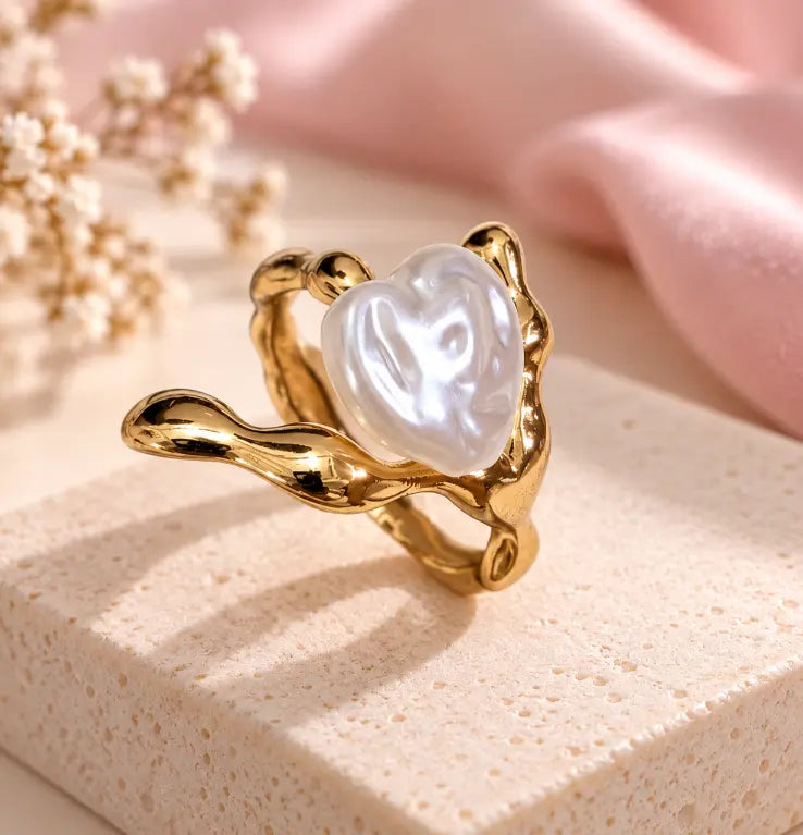 A gold ring with a sculpted, organic-shaped setting holding a lustrous white pearl, displayed on a textured cream block with soft pink fabric and dried flowers in the background.