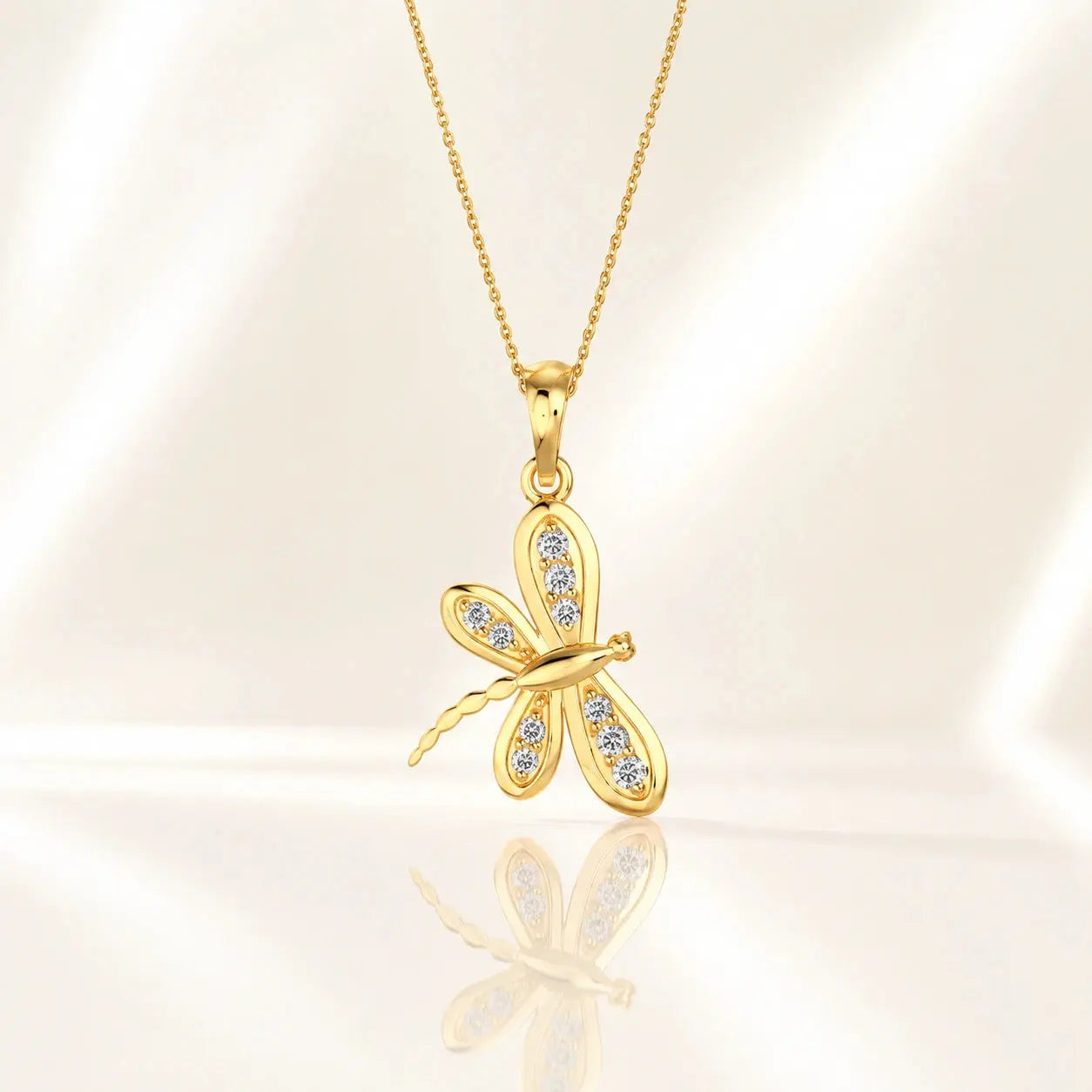 A gold dragonfly pendant necklace adorned with small clear crystals, displayed on a soft, light beige satin background with a subtle reflection below. – Aurelia Dragonfly Lab-Grown Diamond Pendant