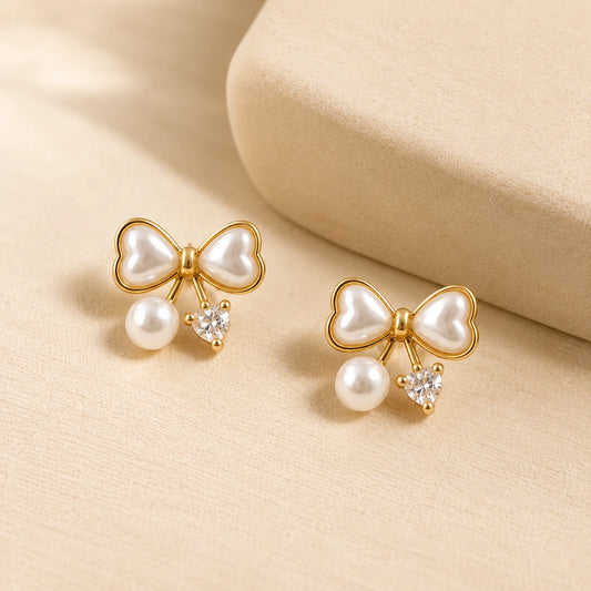 A pair of gold bow stud earrings with white faux pearls and small clear heart-shaped crystals, displayed on a beige textured surface. – Pearl Bow 18K Gold Plated Stud Earrings