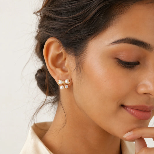 A close-up of a woman wearing a pair of gold stud earrings with white pearl-like beads and small clear stones, shown against a soft light background. – Pearl Bow 18K Gold Plated Stud Earrings
