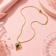 A gold heart-shaped pendant necklace with a green oval center stone, displayed on a soft pink background with decorative fabric and glass props. – Emerald Heart Pendant Necklace