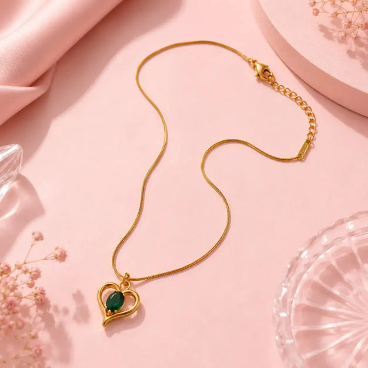 A gold heart-shaped pendant necklace with a green oval center stone, displayed on a soft pink background with decorative fabric and glass props. – Emerald Heart Pendant Necklace