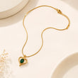 A gold chain necklace with a heart-shaped pendant featuring a dark green oval stone, displayed on a warm off-white background with subtle decorative ceramic shapes and dried flowers. – Emerald Heart Pendant Necklace