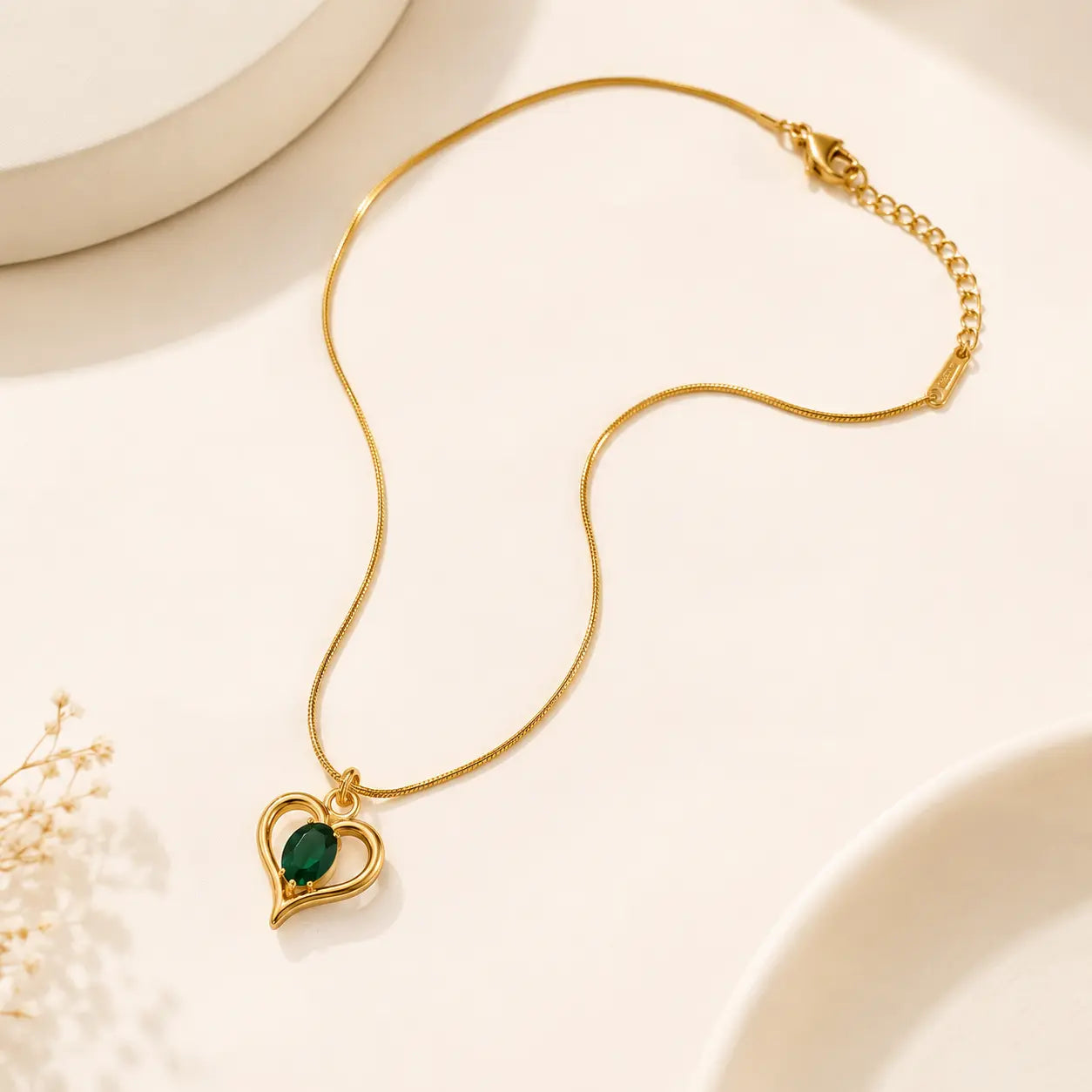 A gold chain necklace with a heart-shaped pendant featuring a dark green oval stone, displayed on a warm off-white background with subtle decorative ceramic shapes and dried flowers. – Emerald Heart Pendant Necklace