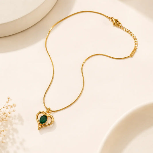 A gold chain necklace with a heart-shaped pendant featuring a dark green oval stone, displayed on a warm off-white background with subtle decorative ceramic shapes and dried flowers. – Emerald Heart Pendant Necklace
