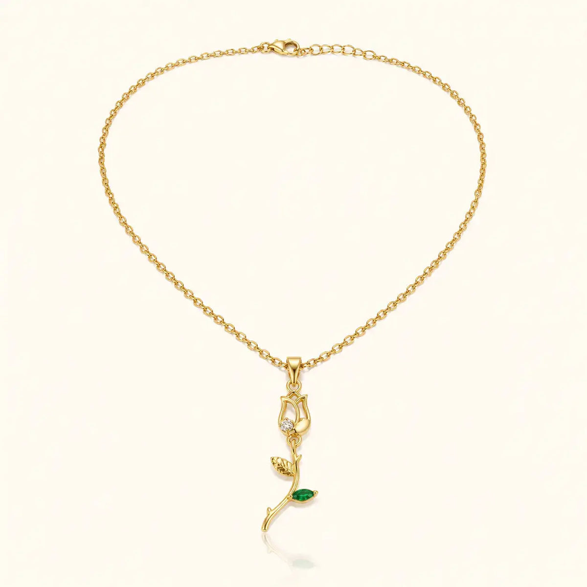 A delicate gold chain necklace with a pendant featuring a small green leaf and a clear crystal, displayed on a light cream background. – Emerald Rose Pendant Necklace
