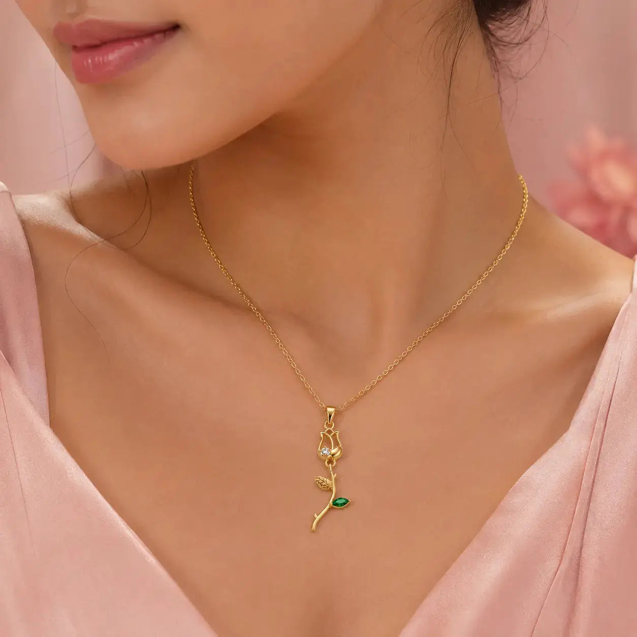 A close-up of a woman wearing a delicate gold necklace with a rose-shaped pendant featuring a small green stone accent, styled with a soft pink satin top. – Emerald Rose Pendant Necklace