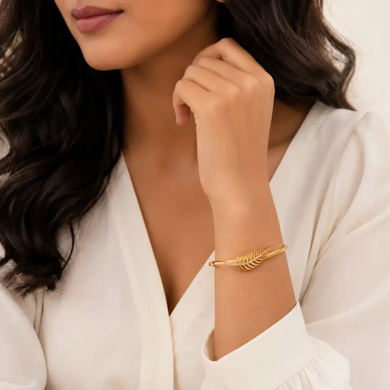 A close-up of a woman wearing a delicate gold leaf-shaped bangle bracelet on her wrist, paired with a white blouse. – Feather Grace Crystal Bracelet – 18K Gold Plated