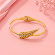 A gold hinged bangle bracelet with a textured leaf design accented by a row of small clear stones, displayed on a soft pink background with decorative pink fabric and dried flowers. – Feather Grace Crystal Bracelet – 18K Gold Plated