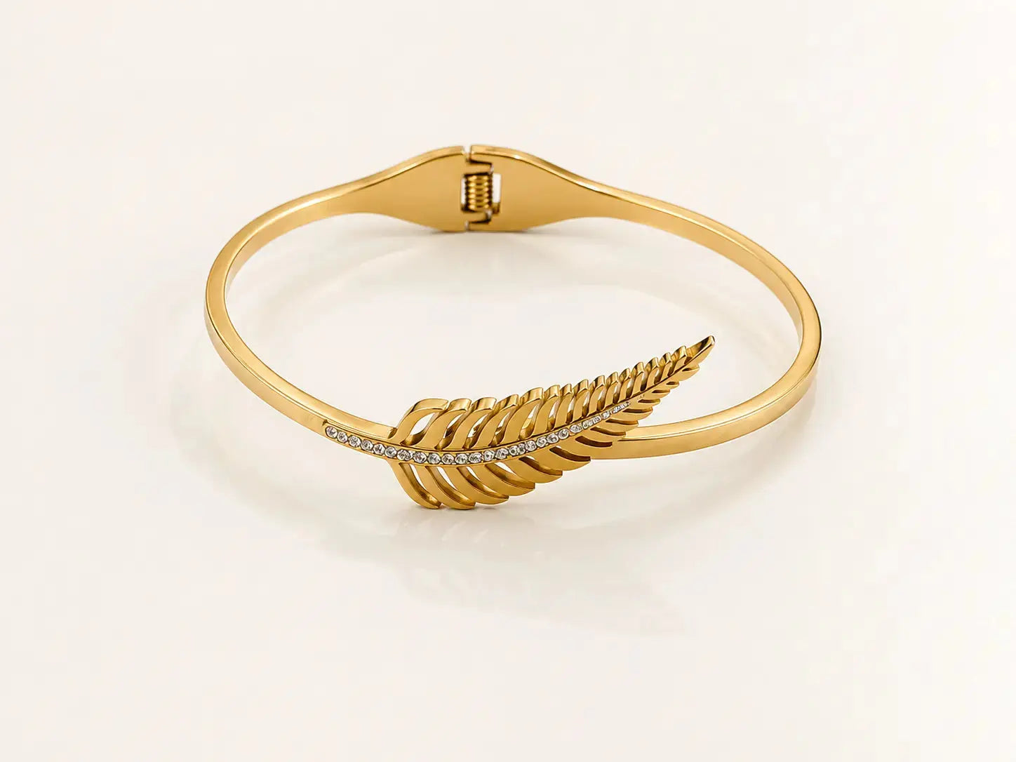 A gold bangle bracelet featuring a detailed silver fern motif set with small clear stones, displayed on a clean white background. – Feather Grace Crystal Bracelet – 18K Gold Plated