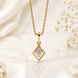 A gold chain necklace with a diamond-shaped pendant featuring a clear pavé center, displayed on a soft white satin fabric background with delicate dried flowers. – Geometric Spark Diamond Pendant Necklace