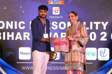 A man in a navy blue shirt and white trousers and a woman in a traditional pink and gold embroidered salwar kameez outfit are standing together on a stage, holding a pink box in front of a blue…
