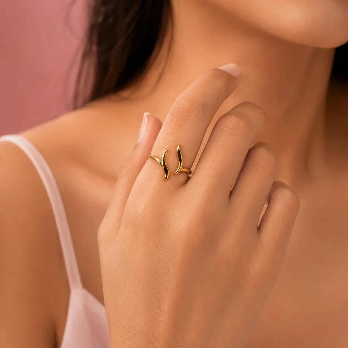 A close-up lifestyle shot of a model's hand wearing a delicate gold ring with two curved leaf-like shapes, set against a soft pink background. – Luna Wave Adjustable Ring