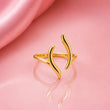 A gold open ring with two curved, leaf-like branches extending upward, displayed on a reflective pink surface with a draped pink fabric in the background. – Luna Wave Adjustable Ring
