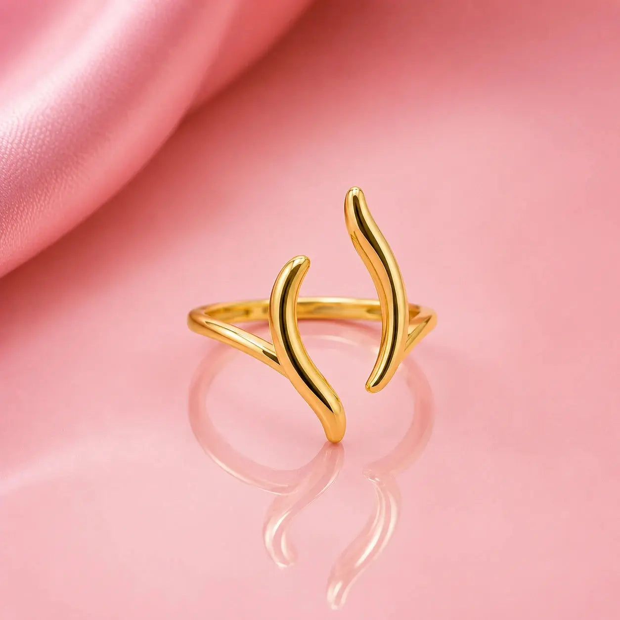 A gold open ring with two curved, leaf-like branches extending upward, displayed on a reflective pink surface with a draped pink fabric in the background. – Luna Wave Adjustable Ring