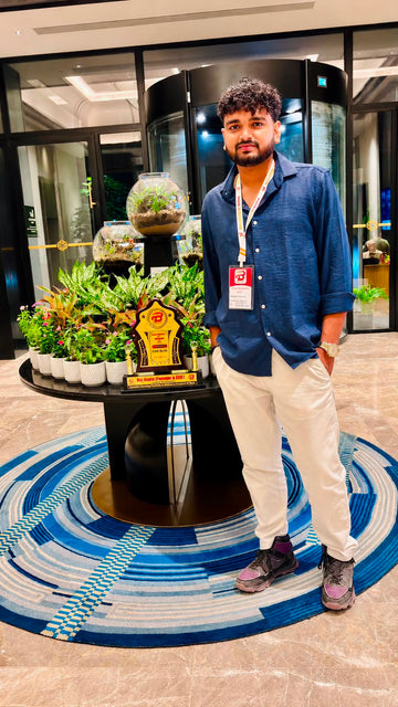 A man wearing a blue button-up shirt, off-white trousers, and dark sneakers stands in a lobby next to a round display table with potted plants and a gold trophy. He is standing on a blue and white…