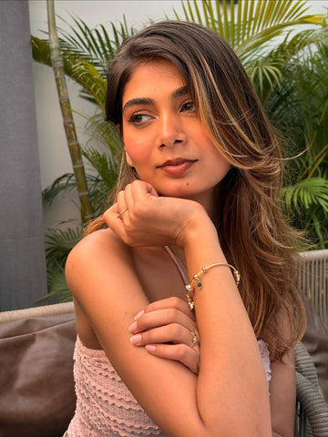 A woman with long brown hair with blonde highlights rests her chin on her hand, wearing a light pink textured top and gold jewelry, including a bracelet and rings, in an outdoor setting with green…