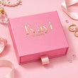 A pink gift box with the brand name 'Tvayi' in gold foil lettering, styled on a pink background with decorative pearls, a perfume bottle, rings, and ribbons.