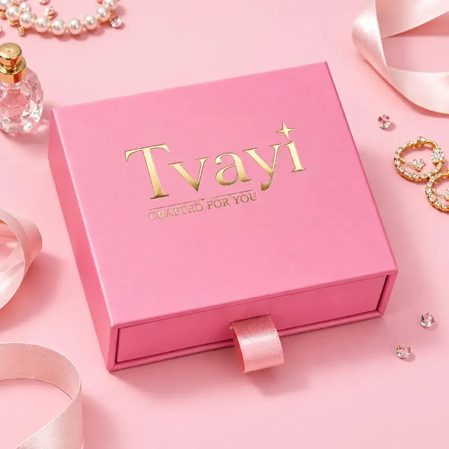 A pink gift box with the brand name 'Tvayi' in gold foil lettering, styled on a pink background with decorative pearls, a perfume bottle, rings, and ribbons.